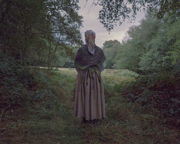 Enclavada en los exuberantes paisajes de Galicia, una mujer vestida con el traje tradicional gallego posa ante una pradera en el límite de un enclave boscoso. La vegetación, testimonio de la histórica abundancia de lluvias en Galicia, se despliega ante ella. La esencia del legado verde y perdurable de Galicia, modelado por las lluvias constantes que han bendecido la región, se encarna en la silueta de esta mujer, en una conexión con la naturaleza que caracteriza al pueblo gallego.
