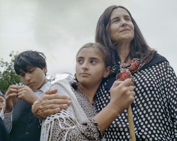 En Palas de Rei, en el corazón de Galicia, un festival anual celebra los valores tradicionales gallegos, donde los vecinos se
										visten con ropa de época y representan piezas teatrales que ponen de relieve los problemas de la Galicia contemporánea. El
										tema de este año fue “la bienvenida”, entendida como el regreso de un emigrante a su tierra gallega. Galicia, históricamente
										una tierra de emigración, marcada por la pobreza endémica y la falta crónica de oportunidades. Muchos emigrantes partieron
										hacia países extranjeros y nunca regresaron —como mis bisabuelos, a quienes nunca conocí—. Otros muchos se fueron
										solo para volver cargando la misma pobreza de la que habían intentado huir. La representación teatral de este año planteó
										“la bienvenida” de forma irónica y satírica: el emigrante regresa, todo el pueblo lo espera con los brazos abiertos creyendo
										que traerá prosperidad, pero vuelve más pobre que nadie, y son los propios vecinos quienes deben ayudarle a sobrevivir. En
										la imagen: una niña que mira hacia atrás mientras una mujer dirige la mirada hacia el futuro. Para mí, esta fotografía es una
										alegoría del tiempo en Galicia, suspendido entre un pasado marcado por la emigración y un futuro incierto.