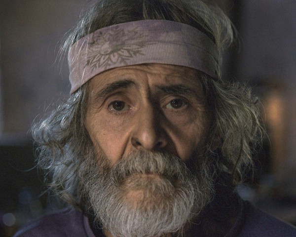 Alfonso, que lleva más de 40 años viviendo en las montañas de Negueira de Muñiz, es uno de los pocos habitantes que quedan
										de la primera comuna hippie establecida más allá del embalse del río Navia. Ha vivido durante más de cuatro décadas
										sin electricidad ni agua corriente. Hornea su propio pan y mantiene una forma de vida casi completamente autosuficiente.
										Alfonso tiene un hijo llamado Isi, de unos treinta años, que vive en una aldea cercana. El camino que conduce al lugar donde
										vive Alfonso sigue sin estar asfaltado. Muchas personas de la zona defendieron durante años su pavimentación, pero
										Alfonso siempre se opuso, afirmando que cuanto más fácil fuera el acceso, más gente llegaría y más se diluiría su forma de
										vida auténtica. Hoy, Alfonso continúa fiel a su manera de vivir: pasa la mayor parte del tiempo leyendo, cuidando su huerto
										y atendiendo a sus animales. Para los jóvenes que hoy habitan las montañas de Negueira, Alfonso es visto casi como una
										especie de sabio o profeta, no solo por el conocimiento acumulado a lo largo de las décadas, sino también por la energía
										personal que irradia su presencia.