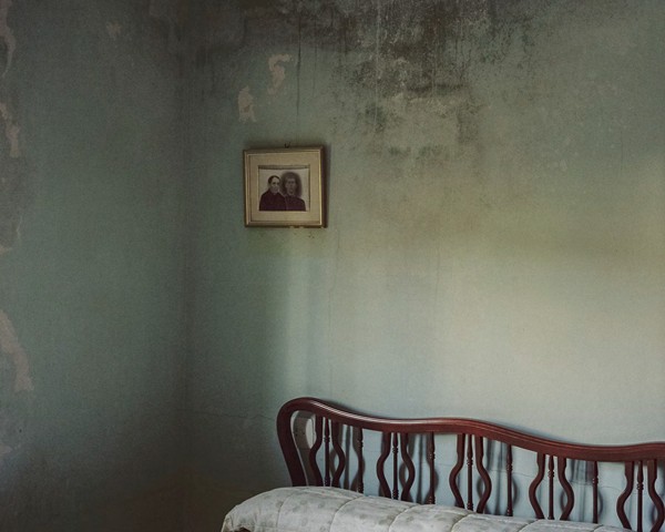 Durante los principales años de la emigración gallega —las décadas de 1970, 1980 y 1990— muchos emigrantes que lograron
										ganar buenas sumas de dinero en el extranjero decidieron construir grandes casas en Galicia, con la esperanza de tener
										algún día un buen lugar donde vivir al regresar. Muchas de estas casas quedaron vacías para siempre, bien porque sus propietarios
										murieron antes de regresar, bien porque finalmente nunca volvieron. En la imagen se aprecia el paso del tiempo y
										el abandono en una pared húmeda de una habitación de una casa construida en la época de la emigración, hoy prácticamente
										abandonada. En la pared cuelga un retrato de los padres de los propietarios de la casa, Claudina y Antonio, que nunca
										llegaron a vivir en esa estancia.
